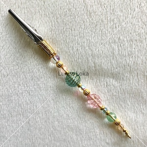 K&ouml;nnte beinhalten: Eine silberfarbene Metall-Krokodilklemme mit einer goldfarbenen Feder und dekorativen Perlen. Die Perlen sind goldfarben, rosa, gr&uuml;n und irisierend. Der Hintergrund zeigt den Text "Luwa Loka".