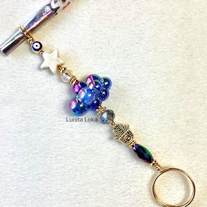 May include: A decorative pen accessory featuring a gold ring, a Hamsa hand charm, and iridescent blue beads. The accessory also includes a white star, an evil eye bead, and the text "Lunità Loka".