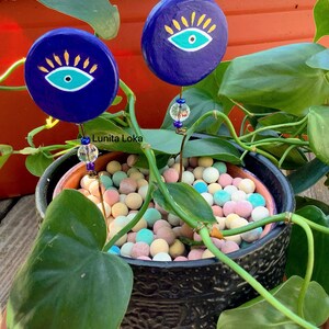 May include: Two dark blue, round plant stakes with an eye design in yellow and teal. The stakes are in a black pot with colorful decorative balls. The plant stakes have a clear bead and wire stem. The text "Lunita Loka" is visible.