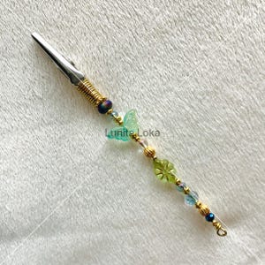K&ouml;nnte beinhalten: Eine silberfarbene Haarspange aus Metall mit einer goldfarbenen Spule und dekorativen Perlen. Die Perlen umfassen einen hellgr&uuml;nen Schmetterling, eine gr&uuml;ne Blume und verschiedene blaue und klare Perlen. Der Text "Lunitia Loka" ist sichtbar.