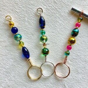 May include: Three colorful beaded keychains with gold, green, blue, and pink beads. Each keychain has a different colored ring at the bottom: gold, silver, and rose gold.