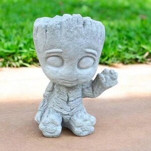 May include: A gray concrete figurine of a character from the Guardians of the Galaxy movie, Groot. The figurine is standing with one arm raised and has a friendly expression.