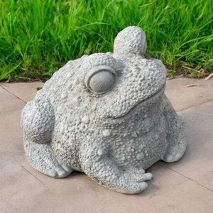 May include: A gray concrete garden statue of a frog with a bumpy texture. The frog is sitting on a gray concrete surface.