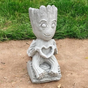 May include: A gray concrete statue of a character from the movie Guardians of the Galaxy, Groot, holding a heart in his hands. The statue is sitting on a base and is designed to be a planter.