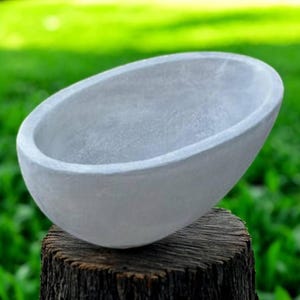 Peut inclure: Un bol en béton gris de forme ovale. Le bol est posé sur une souche de bois vieillie sur un fond vert flou. La surface lisse du bol contraste avec la texture rugueuse du bois.