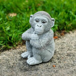 May include: A small gray concrete statue of a monkey sitting with its hands clasped in front of it. The monkey has a realistic face with large eyes and a small mouth.