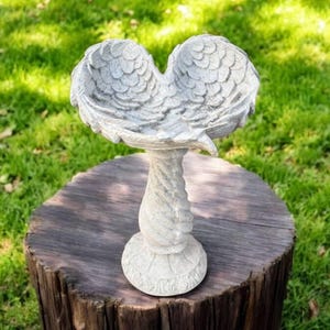 May include: A white ceramic bird bath shaped like a pair of angel wings. The bath is perched on a pedestal with a swirling design.
