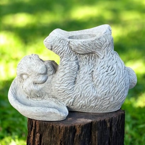 May include: A light grey concrete bunny planter, sitting on a weathered wooden stump. The bunny is sculpted with detailed fur texture, featuring a large opening for planting. The bunny is in a seated position with its ears back.