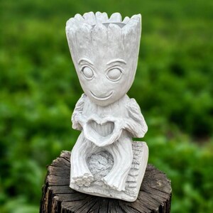 May include: A gray, resin Groot planter, featuring a smiling face and holding a heart-shaped cutout. The planter is designed to sit on a flat surface and has a textured, bark-like appearance. The planter is approximately 8 inches tall.
