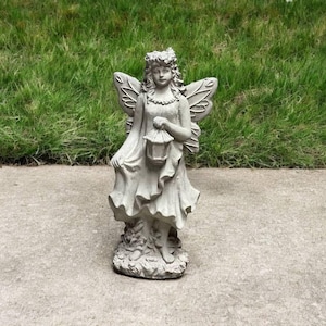 May include: A gray concrete fairy statue with wings, holding a lantern. The fairy is wearing a dress and has a crown of leaves on her head.