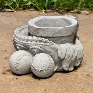 May include: A gray concrete planter shaped like a baseball glove holding two baseballs. The planter has a small pot inside for planting.