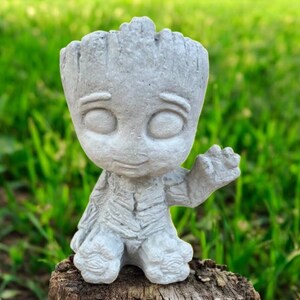 May include: A gray concrete planter shaped like the character Groot from Guardians of the Galaxy. The planter has a detailed textured surface and is posed with one arm raised in a wave. The planter is sitting on a wooden stump.