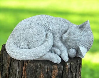 May include: A gray cat sculpture in a curled sleeping position. The cat is resting on a weathered tree stump, with its tail curled around its body. The background is a blurred green.