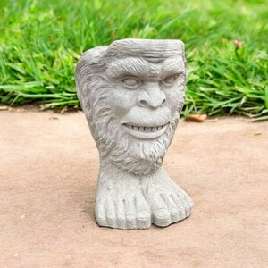 May include: A gray concrete garden statue of a creature with a human-like face and feet. The creature has a wide mouth and a beard.