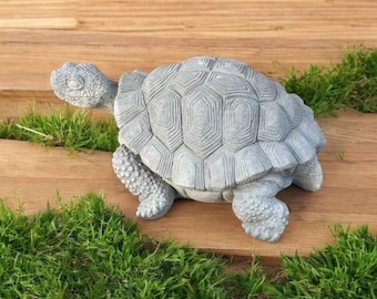 May include: A gray concrete turtle statue with a detailed shell design. The turtle is sitting on a wooden surface with green grass around it.