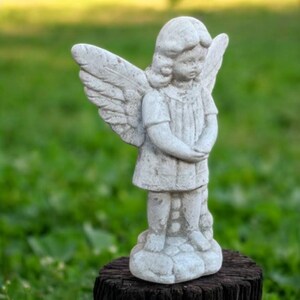 May include: A weathered, gray stone angel statue with outstretched wings. The angel is depicted in a child-like form, wearing a short-sleeved tunic and standing on a textured base. The statue is placed outdoors, with a blurred green background.