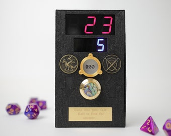 Può includere: Una scatola nera con un display digitale che mostra i numeri 23 e 5. La scatola ha un simbolo di dado D20 dorato e un piccolo pulsante dorato con un intarsio di madreperla. Il testo "Cunr into your fate. Roll to find the answer..." è stampato sulla scatola. Ci sono diversi dadi viola sparsi su una superficie bianca davanti alla scatola.