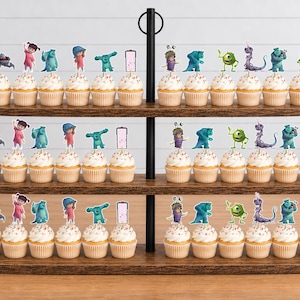 May include: A three-tiered wooden cupcake stand holding numerous cupcakes topped with colourful character paper toppers. The toppers feature various animated figures including a green one-eyed creature, a blue furry monster, and a small child.