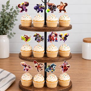 May include: A three-tiered wooden cupcake stand holding twelve cupcakes, each topped with a white frosting swirl, colorful sprinkles, and a paper cutout of a Donkey Kong character. The stand features a black metal central rod and handle.