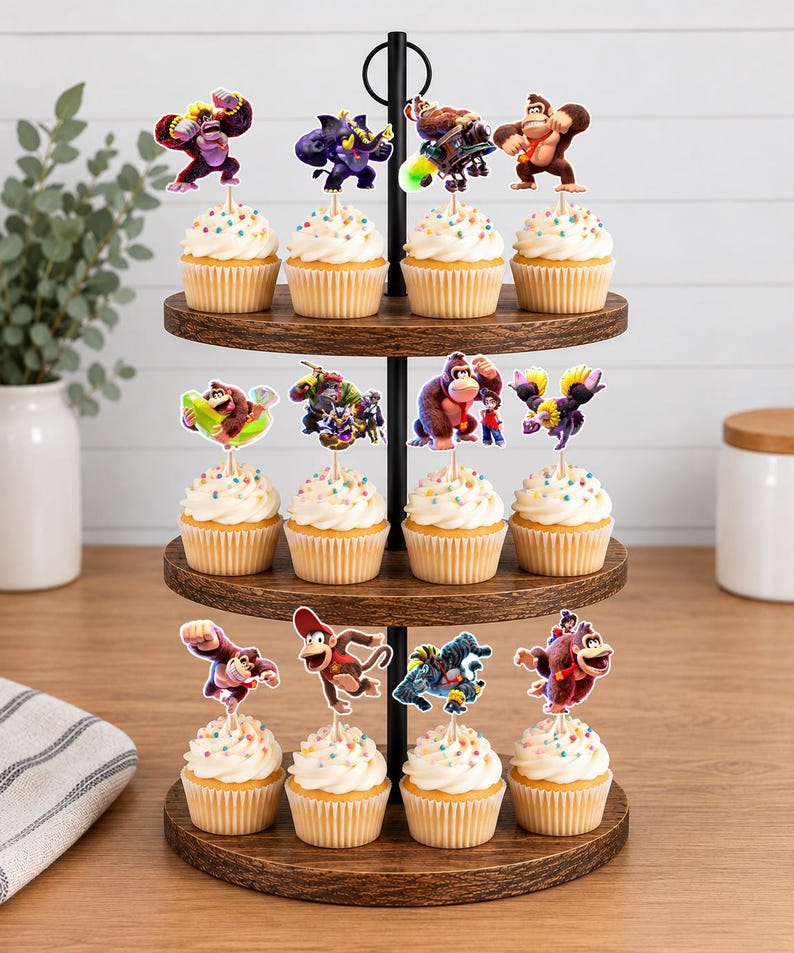 May include: A three-tiered wooden cupcake stand holding twelve cupcakes, each topped with a white frosting swirl, colorful sprinkles, and a paper cutout of a Donkey Kong character. The stand features a black metal central rod and handle.