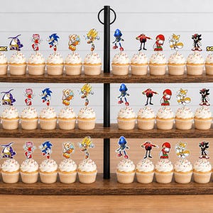 May include: A three-tiered wooden cupcake stand holding numerous cupcakes topped with colorful paper cutouts of Sonic the Hedgehog characters including Sonic, Tails, Knuckles, Shadow, Amy, and Dr. Eggman, with the word SONIC printed on the stand.