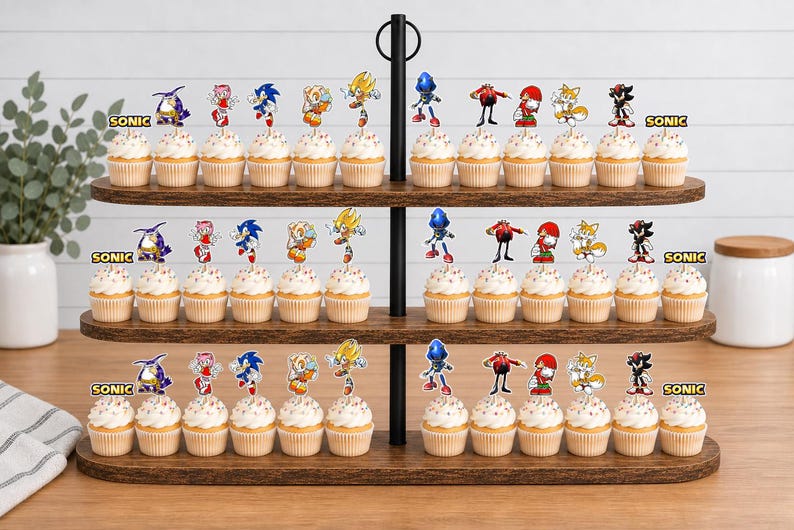 May include: A three-tiered wooden cupcake stand holding numerous cupcakes topped with colorful paper cutouts of Sonic the Hedgehog characters including Sonic, Tails, Knuckles, Shadow, Amy, and Dr. Eggman, with the word SONIC printed on the stand.