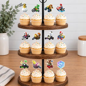 Op de afbeelding: Een houten cupcake-standaard met drie niveaus en twaalf cupcakes, bedekt met witte glazuur en kleurrijke hagelslag. Elke cupcake is voorzien van een decoratieve papieren topper met diverse Mario Kart-personages en items, waaronder racers op voertuigen.
