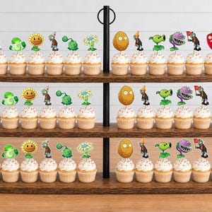 May include: A three-tiered wooden cupcake stand holding numerous cupcakes topped with decorative paper cutouts featuring various characters from the Plants vs. Zombies game, including sunflowers, walnuts, peashooters, and various zombie figures.