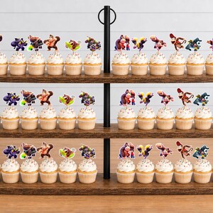 May include: A three-tiered wooden cupcake stand holding numerous cupcakes topped with colorful Donkey Kong character paper toppers. The stand features a central black metal pole with a ring handle at the top, displaying a collection of themed party treats.