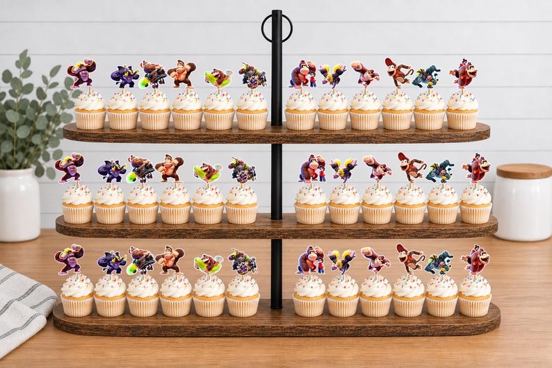 May include: A three-tiered wooden cupcake stand holding numerous cupcakes topped with colorful Donkey Kong character paper toppers. The stand features a central black metal pole with a ring handle at the top, displaying a collection of themed party treats.