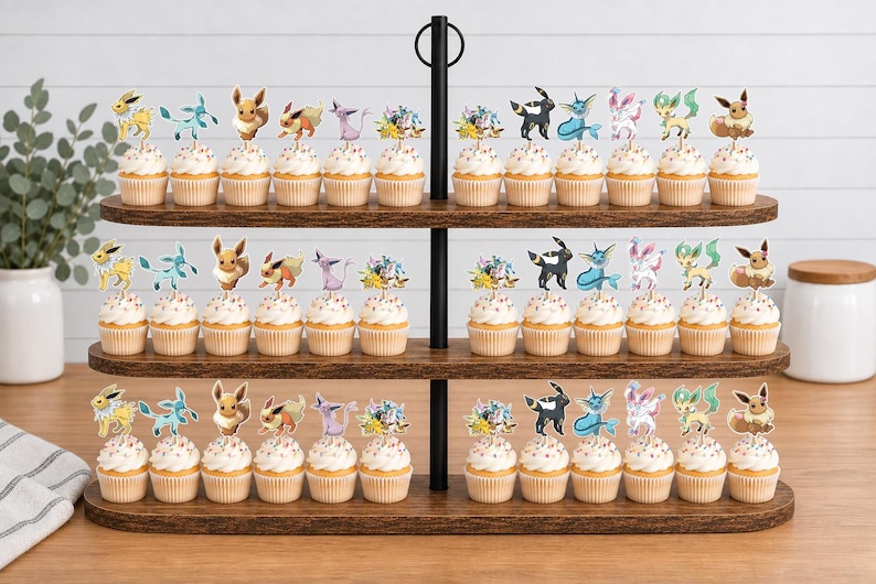 May include: A three-tiered wooden cupcake stand holding numerous cupcakes, each topped with a decorative paper cutout of various Eevee evolution characters. The stand features a central black metal pole and a dark wood grain finish on the shelves.