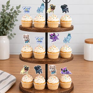 Puede incluir: Un soporte de madera de tres niveles con doce cupcakes cubiertos de glaseado blanco y chispas de colores. Cada cupcake tiene un adorno de papel con personajes y el texto Puppy Dog Pals de la serie de animaci&oacute;n.