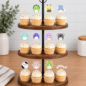May include: A three-tiered wooden cupcake stand holding twelve cupcakes topped with colorful sprinkles and various Squishmallows character paper toppers, including a bee, a dinosaur, a fox, a koala, a cow, and the Squishmallows logo text.