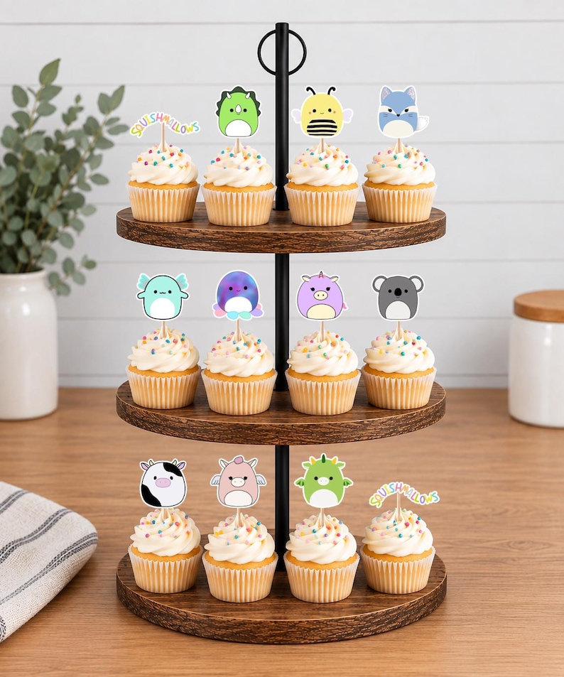 May include: A three-tiered wooden cupcake stand holding twelve cupcakes topped with colorful sprinkles and various Squishmallows character paper toppers, including a bee, a dinosaur, a fox, a koala, a cow, and the Squishmallows logo text.