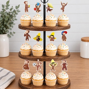 May include: A three-tiered wooden cupcake stand holding twelve cupcakes topped with Curious George character paper toppers. Each topper features the brown monkey in various poses, including holding a banana, a balloon, a kite, a soccer ball, or wearing a yellow hat.