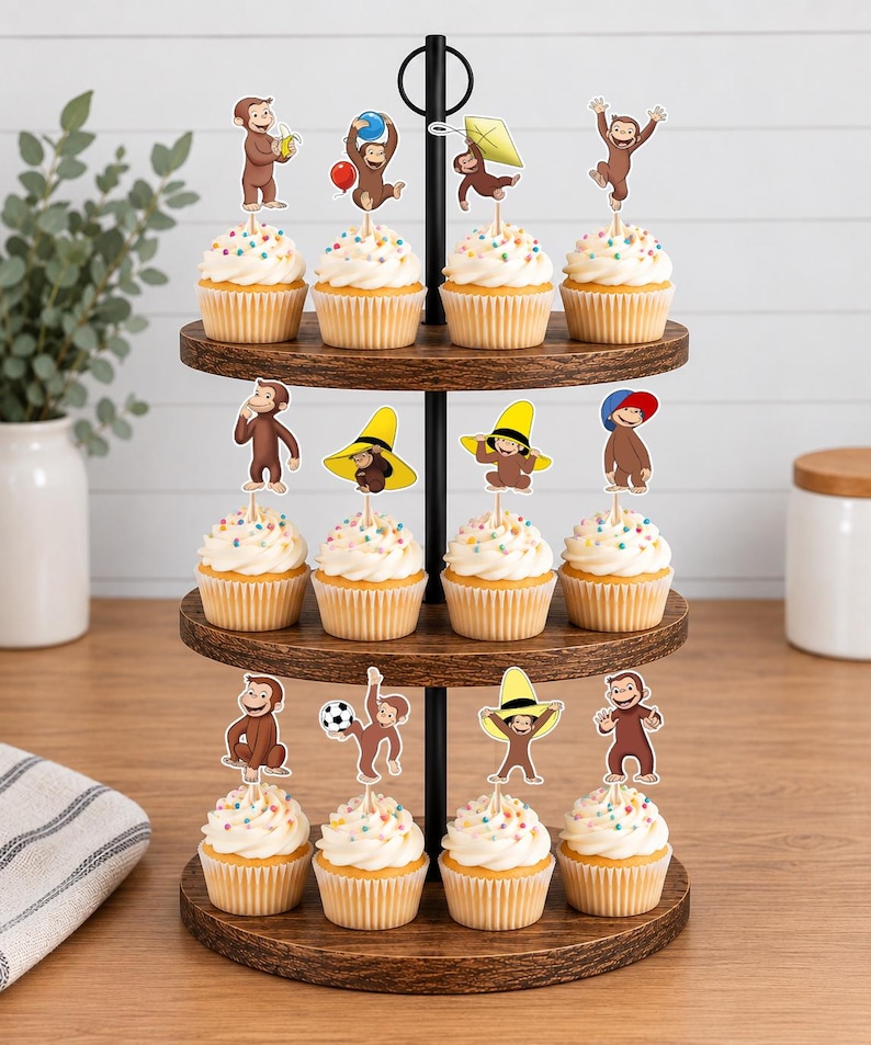 May include: A three-tiered wooden cupcake stand holding twelve cupcakes topped with Curious George character paper toppers. Each topper features the brown monkey in various poses, including holding a banana, a balloon, a kite, a soccer ball, or wearing a yellow hat.