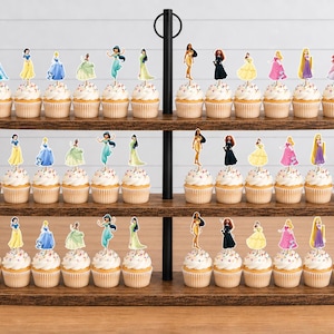 May include: A three-tiered wooden cupcake stand holding numerous cupcakes, each topped with a small, flat, printed paper cutout of a different female character in a colorful dress. The stand features a black metal central support rod with a ring handle.