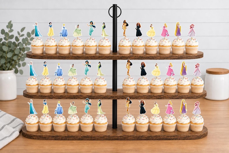 May include: A three-tiered wooden cupcake stand holding numerous cupcakes, each topped with a small, flat, printed paper cutout of a different female character in a colorful dress. The stand features a black metal central support rod with a ring handle.