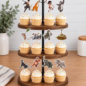 May include: A three-tiered wooden cupcake stand holding twelve cupcakes, each topped with a white frosting swirl and colorful sprinkles. Each cupcake features a decorative paper topper depicting various animated characters from The Jungle Book.