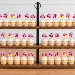 May include: A three-tiered wooden cupcake stand holding numerous cupcakes topped with pink Kirby character toppers. Each cupcake features a light-colored frosting and a small, colorful paper cutout of the video game character Kirby in various poses.
