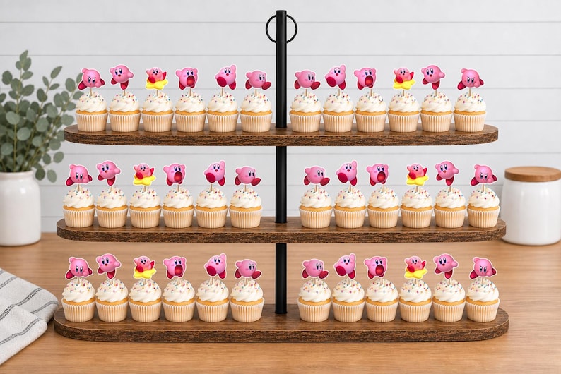 May include: A three-tiered wooden cupcake stand holding numerous cupcakes topped with pink Kirby character toppers. Each cupcake features a light-colored frosting and a small, colorful paper cutout of the video game character Kirby in various poses.