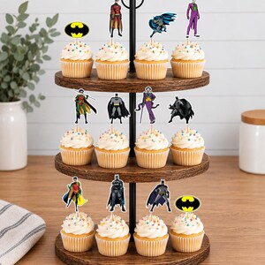 May include: A three-tiered wooden cupcake stand holding twelve cupcakes topped with white frosting and colorful sprinkles. Each cupcake features a decorative paper topper depicting various Batman-themed characters, including Batman, Robin, the Joker, and the yellow bat logo.