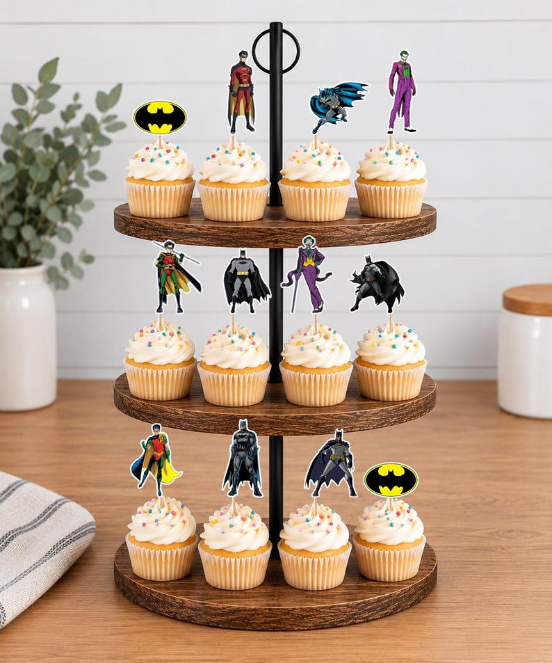 May include: A three-tiered wooden cupcake stand holding twelve cupcakes topped with white frosting and colorful sprinkles. Each cupcake features a decorative paper topper depicting various Batman-themed characters, including Batman, Robin, the Joker, and the yellow bat logo.