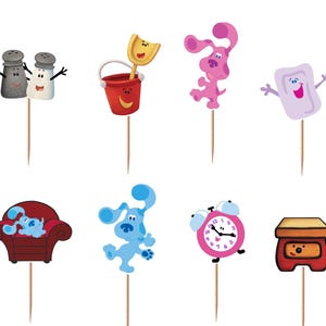 May include: A set of twelve decorative cupcake toppers featuring colorful cartoon characters and objects from Blue's Clues, including blue and magenta dogs, a red bucket, a mailbox, a pink alarm clock, a red armchair, and a soap bar on wooden picks.
