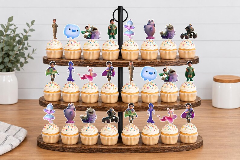 May include: A three-tiered wooden cupcake stand holding twenty-four cupcakes, each topped with a colourful character cutout. The toppers feature various animated figures, including small humanoids, blue blobs, and purple creatures, arranged in rows.