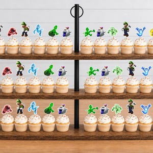 Puede incluir: Un soporte de madera de tres niveles con numerosos cupcakes decorados con adornos de papel que muestran varios personajes de dibujos animados en verde, azul, rojo y blanco, incluyendo figuras con overol verde y diversos seres fantasmales.