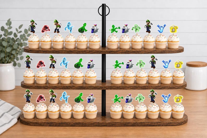 Puede incluir: Un soporte de madera de tres niveles con numerosos cupcakes decorados con adornos de papel que muestran varios personajes de dibujos animados en verde, azul, rojo y blanco, incluyendo figuras con overol verde y diversos seres fantasmales.
