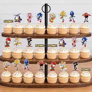 May include: A three-tiered wooden stand holding twenty-four cupcakes topped with colorful paper cutouts of Sonic the Hedgehog characters. Each cupcake features a character topper, and some include the yellow text SONIC on the paper cutout.