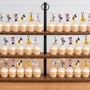 May include: A three-tiered wooden cupcake stand holding numerous cupcakes topped with colorful paper cutouts of various cartoon characters, including Mickey Mouse, Minnie Mouse, Donald Duck, Goofy, Pluto, and Daisy Duck, arranged in rows.