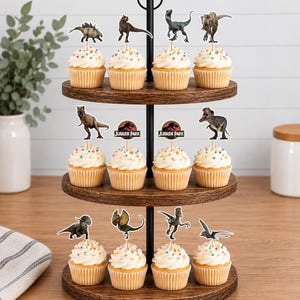 Pu&ograve; includere: Un'alzata in legno a tre ripiani con dodici cupcake guarniti con glassa bianca, zuccherini colorati e vari topper in carta a tema dinosauri, inclusi due loghi circolari di Jurassic Park montati su stecchini di legno.
